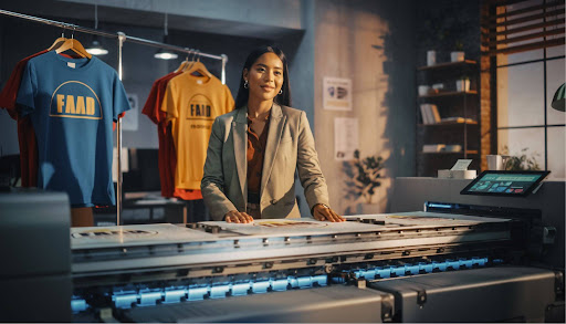 Woman operating a large-format printer in a print shop, with custom t-shirts hanging behind her and a workspace filled with printing equipment.